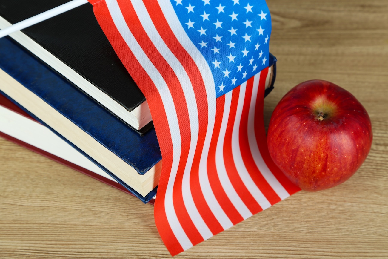 American flag atop a stack of books, an apple to the right, view from above, American spelling versus british